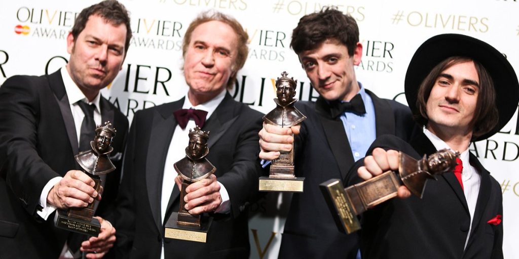 Two men in formal attire hold Olivier Awards trophies, smiling against a branded backdrop.