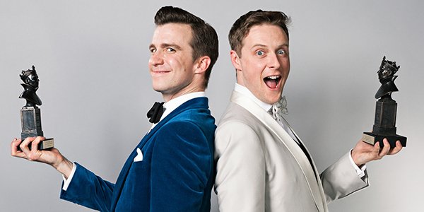 Two men stand back-to-back, one in a blue velvet suit and the other in a white tuxedo, smiling.