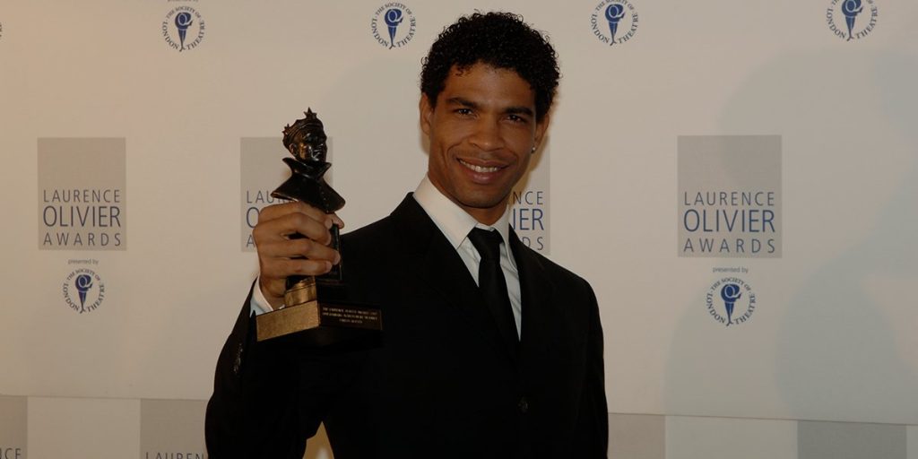 A man in a black suit holds an Olivier Award trophy, smiling against a branded backdrop.