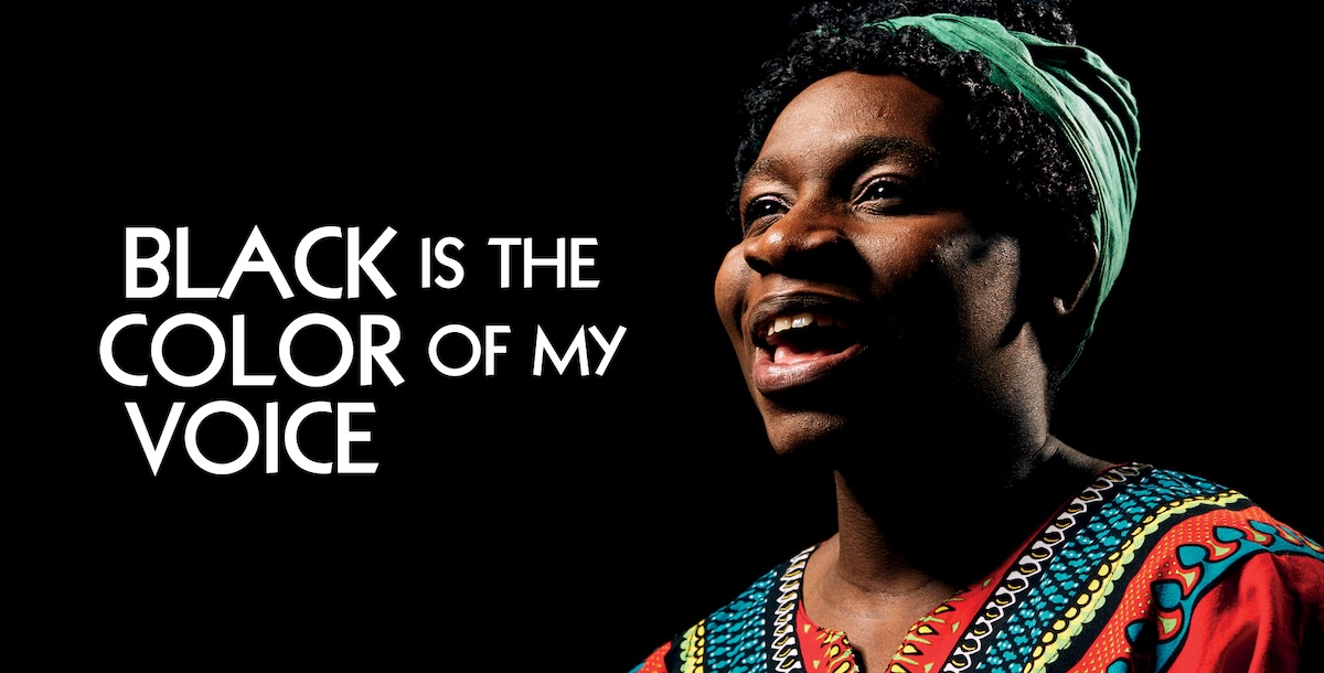 A performer stands on stage, singing passionately, wearing a black outfit, with a spotlight illuminating them. On the right it reads 'Black Is The Color Of My Voice'