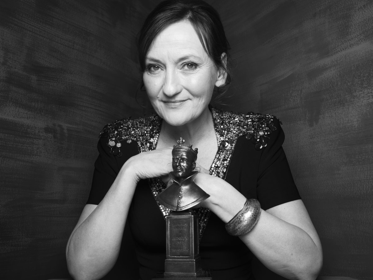 A woman with her elbows leaning on the table in front of her. She has a soft smile. There is an Olivier Award statue on the table in front of her.
