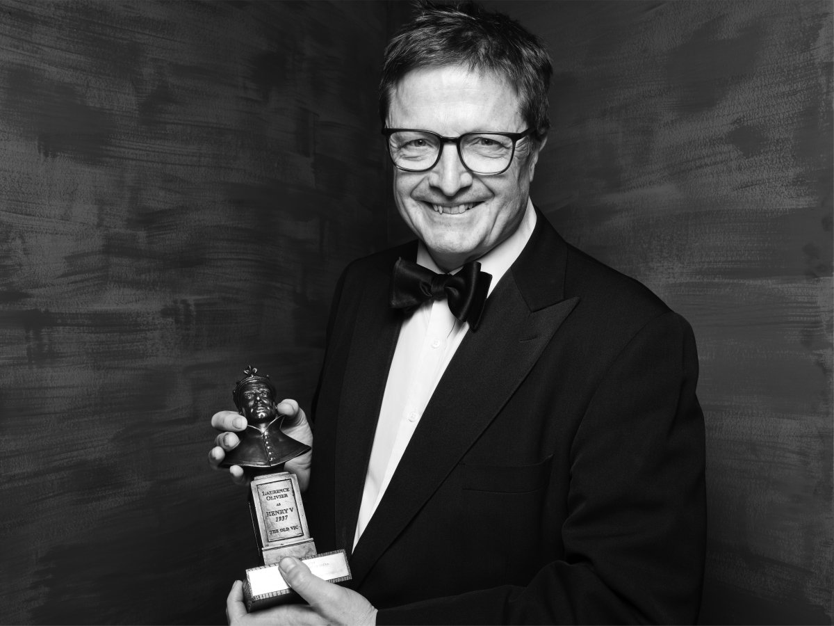 A man wearing glasses holding an Olivier Award in both hands and smiling at the camera.