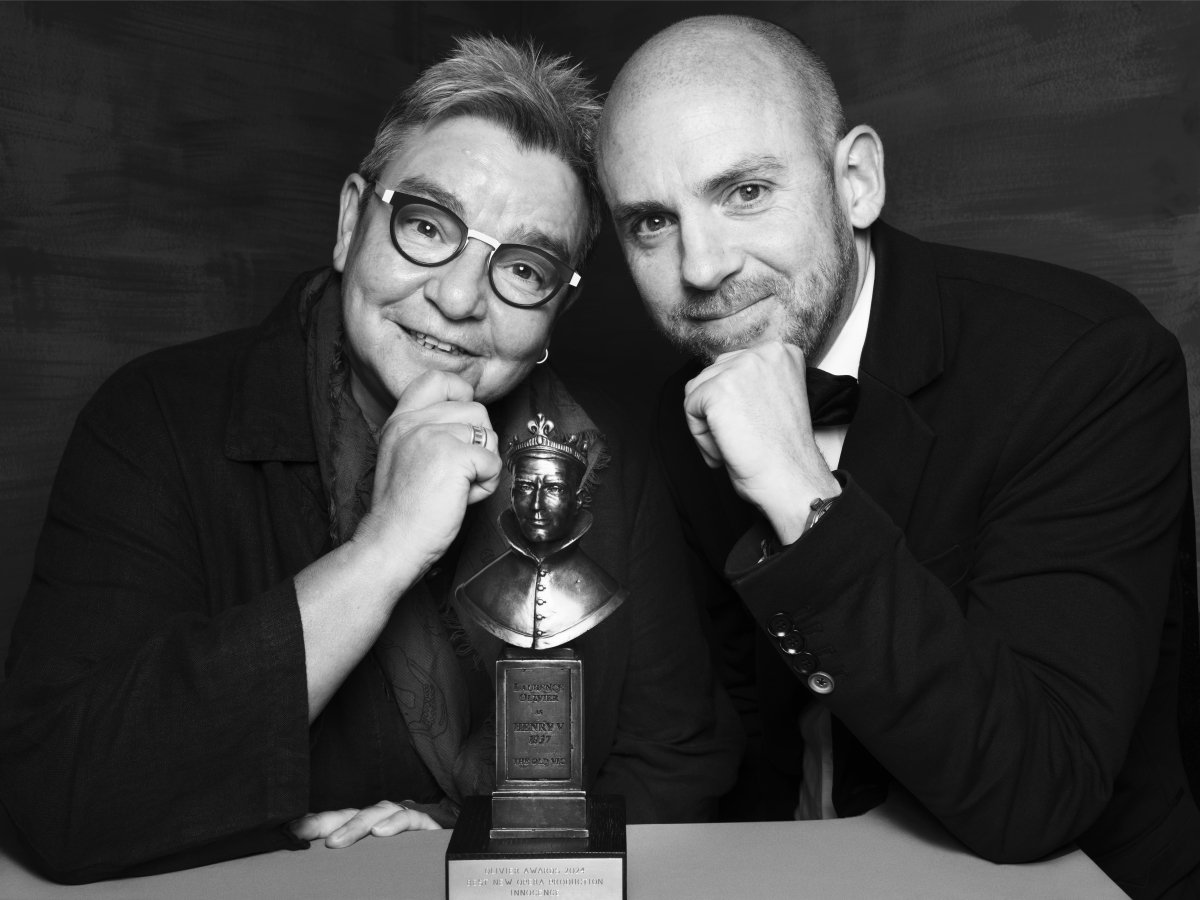 A woman and man smiling at the camera. They both have an elbow on the table and holding a hand up to their chin. There is an Olivier Award statue on the table between them.