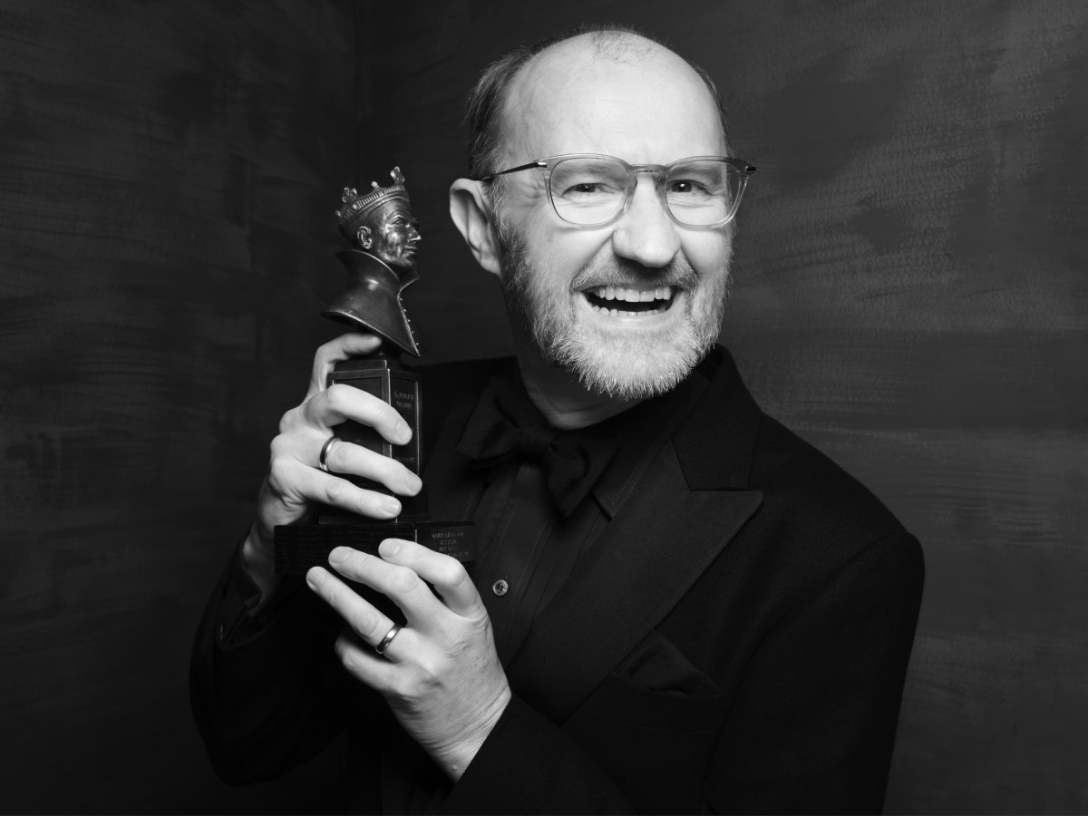 Black and white image of a person smiling to camera, wearing glasses and holding an award.