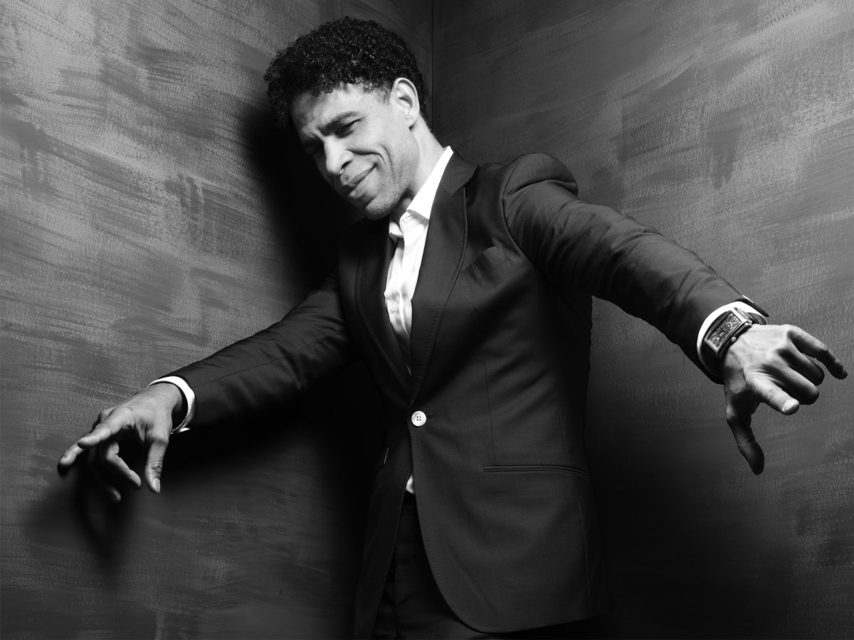 A man in a suit. He is standing in a corner leaning against the wall. His arms are out and he is looking towards the ground.