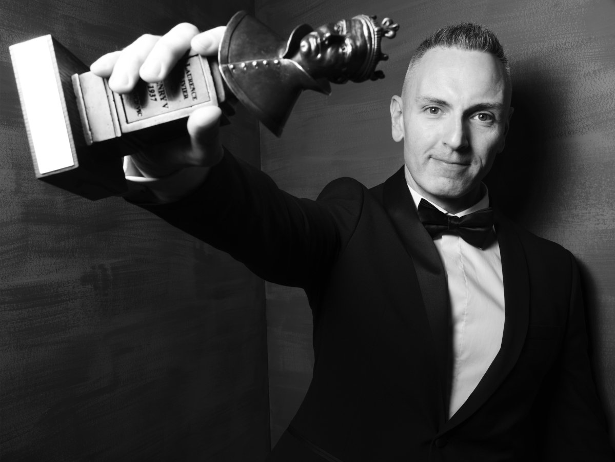 A man in a suit and bow tie. He has a soft smile. He is holding an Olivier Award out in front of him and it is tilted to the side.