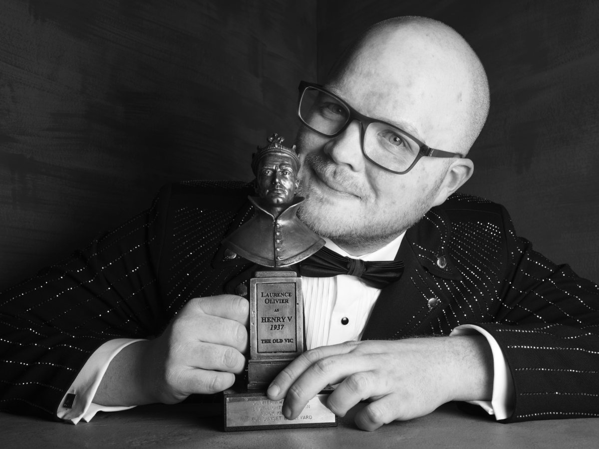 A man with glasses has his arms bent in front of him on a table. There is an Olivier Award statue on the table. The man is resting his chin on the statue, with his head tilted sideways. He has a soft smile.