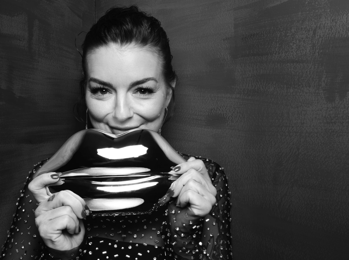 A woman smiling at the camera. She is holding a metal statue of lips under her face.