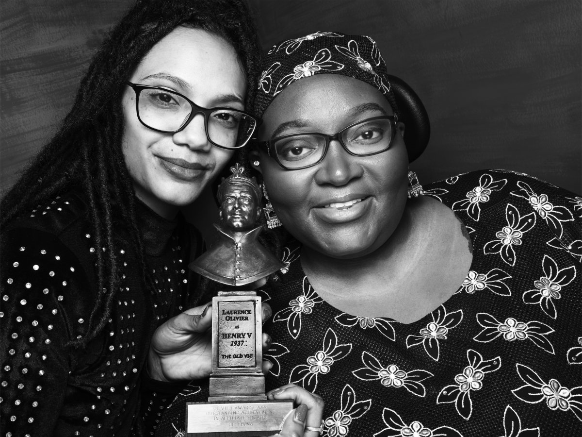 Two women smiling with their heads resting against each other. They are both wearing glasses. They are holding an Olivier Award statue between their faces.