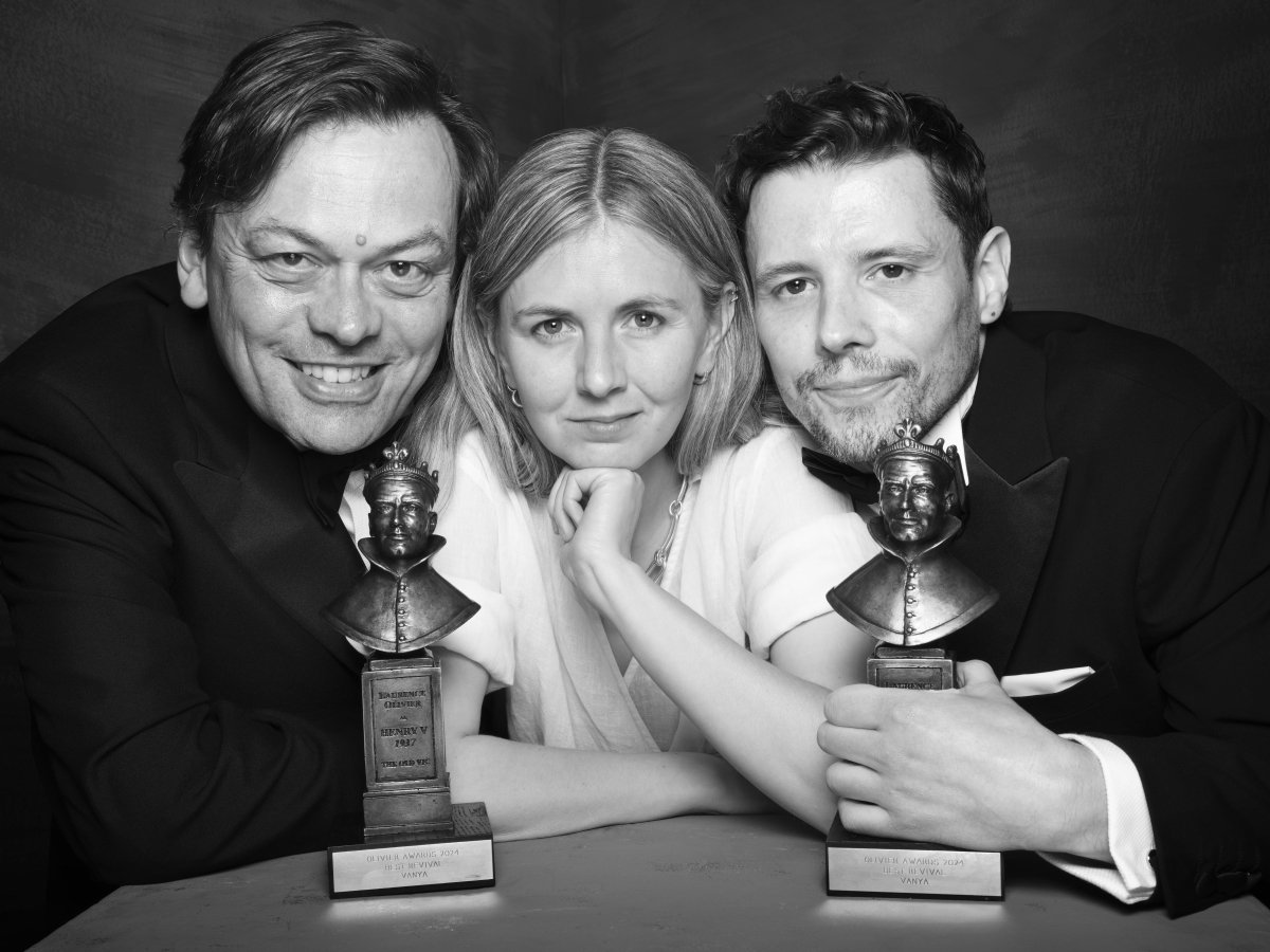 A man, woman and man pose together. The woman smiles with her hand under her chin. The two men are leaning their heads on the woman's shoulders. There are two Olivier Award statues on the table.
