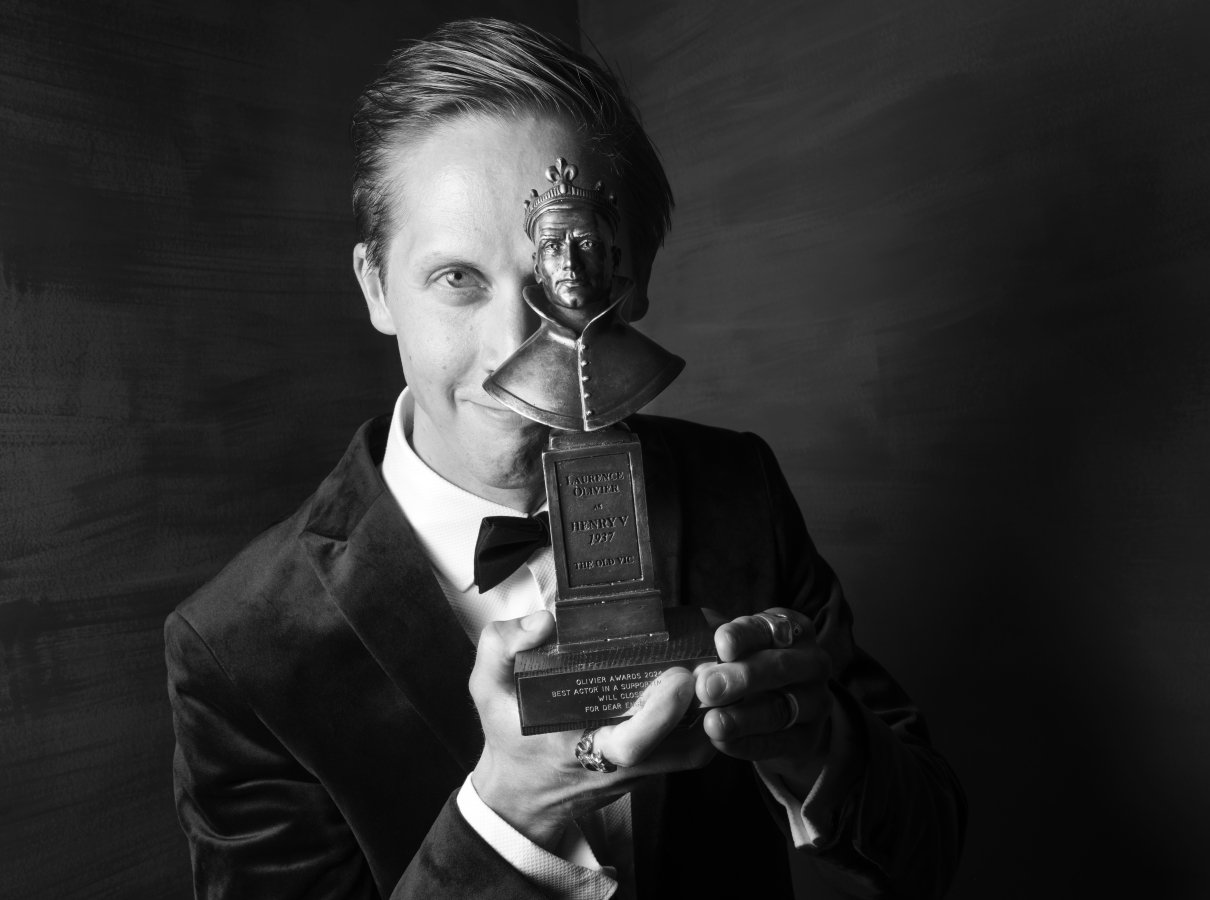 Will Close holding an Olivier Award statue in front of one eye. He is smiling.