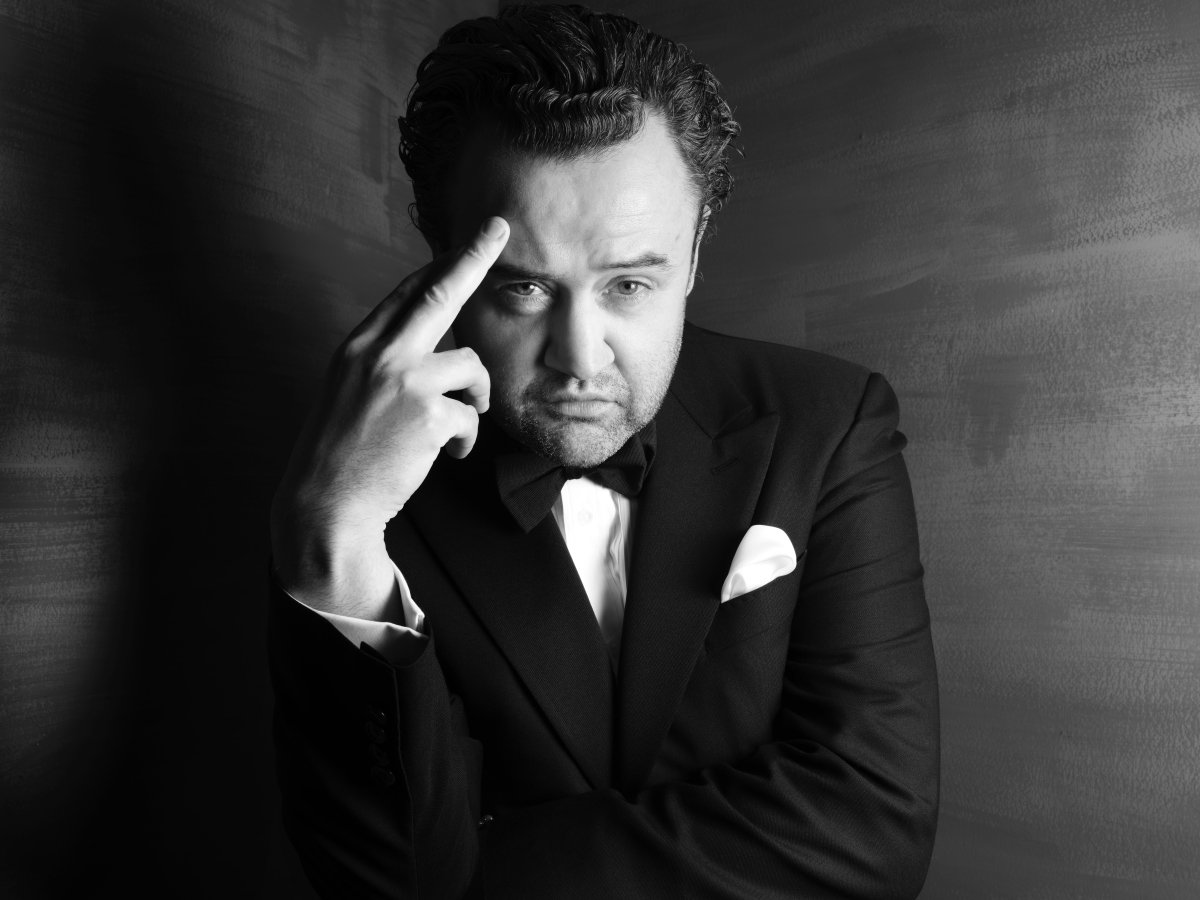 A man in a suit looking seriously at the camera. He has a finger on his forehead.