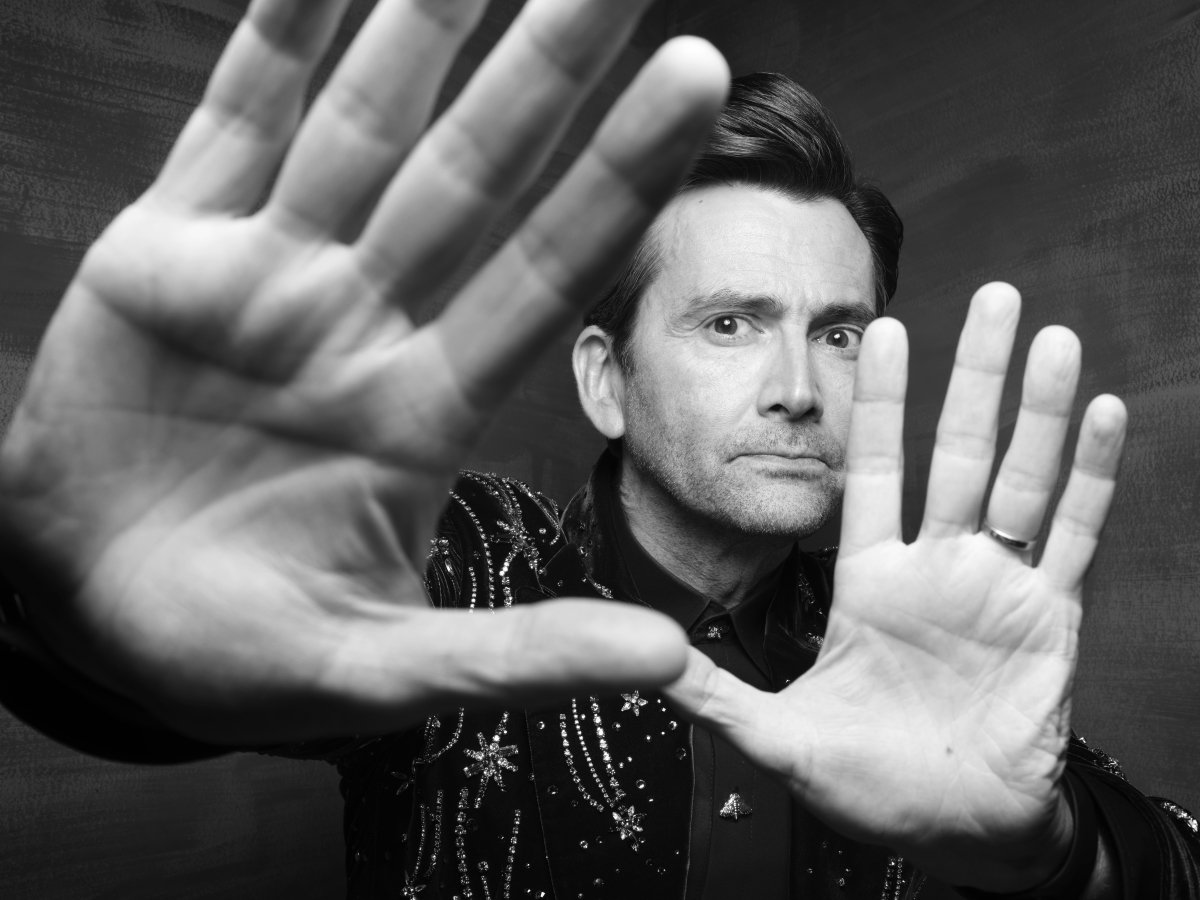 A man in a sequinned suit. He is looking at the camera. He has two hands up towards the camera and you can see his face between them.