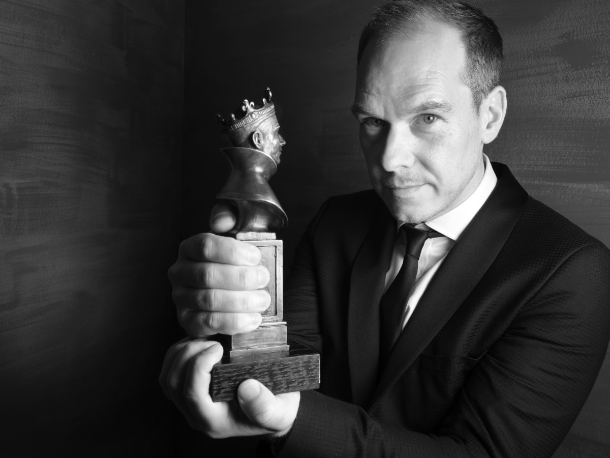 A man in a suit looking at the camera. He is holding an Olivier Award up towards the camera.