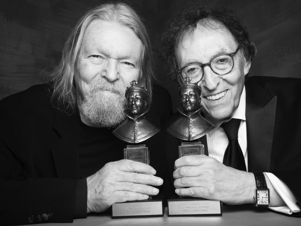 Two people smile at the camera holding awards. Both are dressed in suits and the right is wearing glasses.