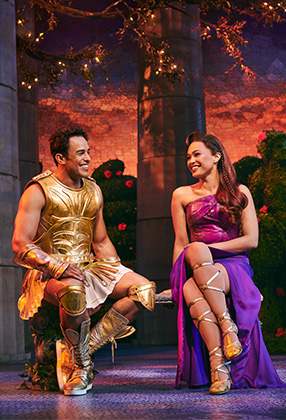 A man in golden armour and a woman in a purple dress sit smiling, surrounded by greenery.