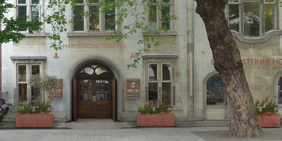 A modern theatre interior with red seating, stage lights, and a large screen displaying "Marylebone Theatre.
