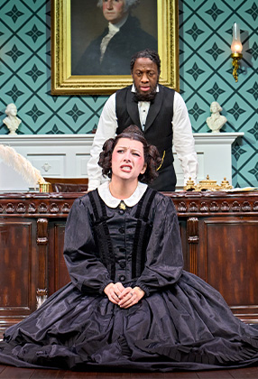 A woman in a black dress sits on the floor, while a man in formal attire stands behind her.
