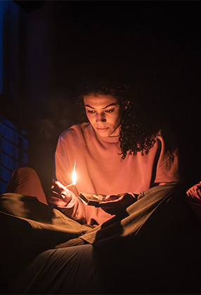 A young woman in a pink top sits cross-legged, holding a candle and looking thoughtfully at it.