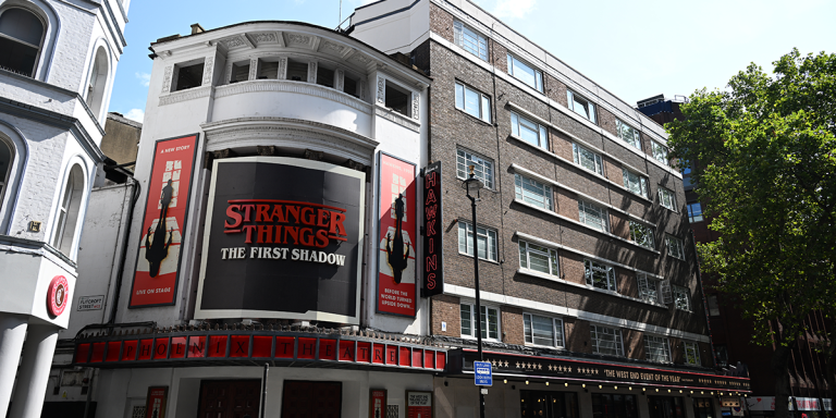 A dark silhouette of a person stands against a bright, textured theatre backdrop.